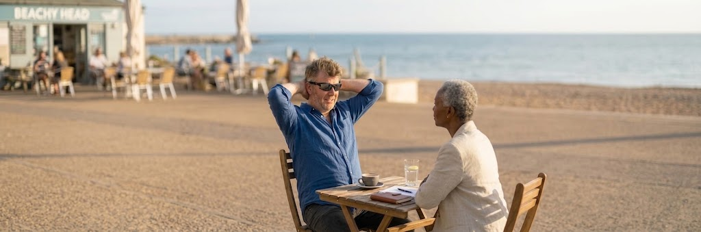 Cameron Dean, creator of GEN:REF, in conversation with an interviewer at a seaside café.